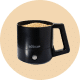 A black, battery-powered milk frother with an ergonomic handle, containing frothed milk, on a light beige background.