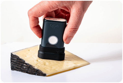 A hand holds a device labeled "Oliso" to vacuum seal a wedge of cheese on a white surface.