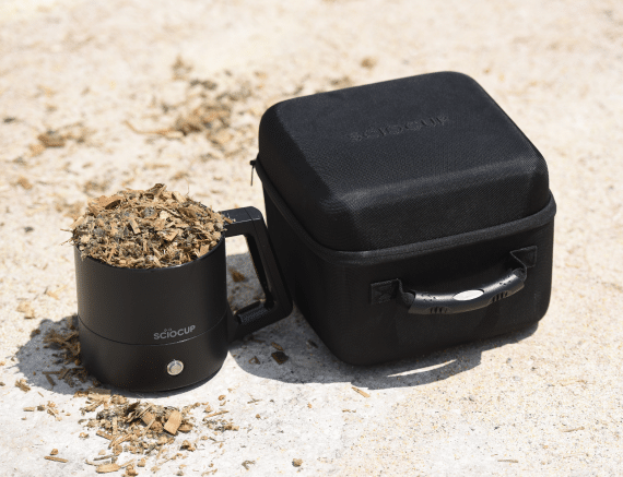 A black SciOcup container filled with dried leaves is next to a black carrying case on a sandy surface.