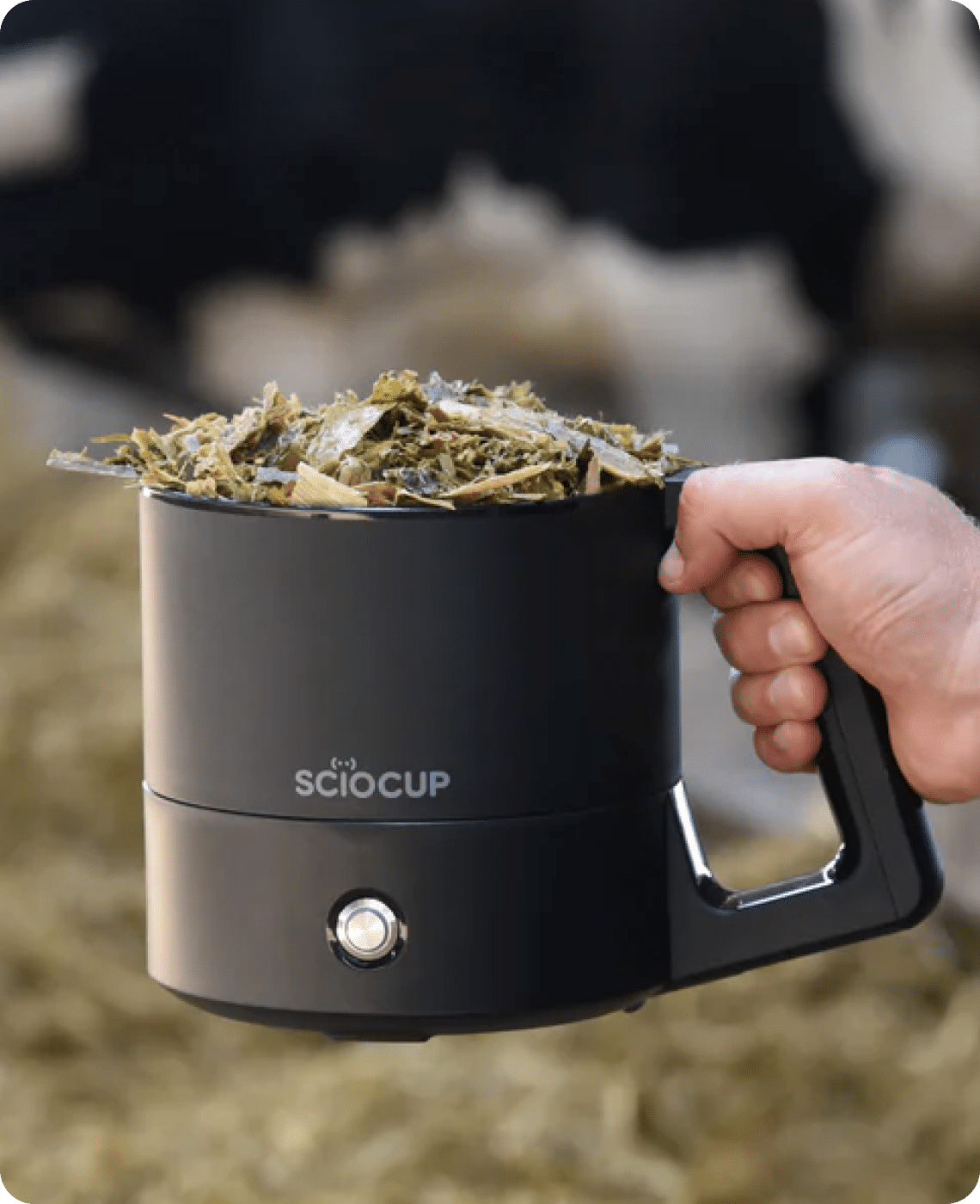 A hand holds a black device labeled "SCIO CUP," filled with chopped plant material, with hay in the background.