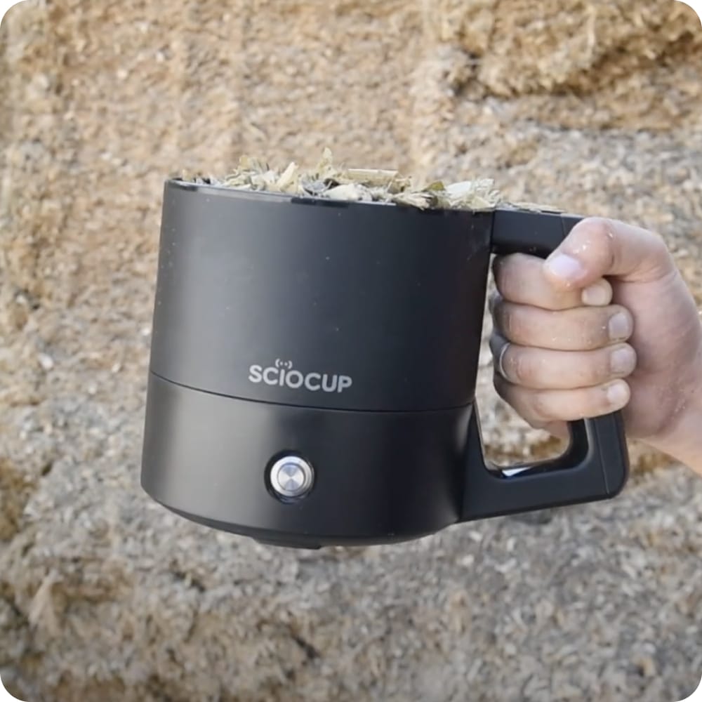 A person holds a black Sciocup filled with soil or organic material against a textured background.