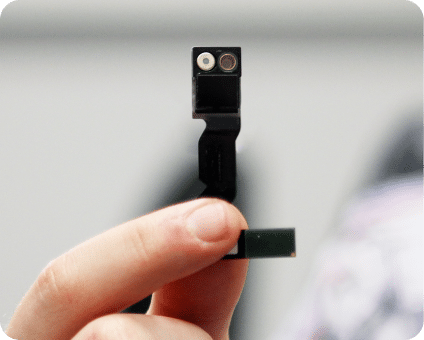 A hand holds a small electronic camera module with dual lenses against a blurred background.
