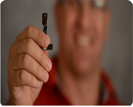 A person holds a small electronic component close to the camera, showing it distinctly while smiling.